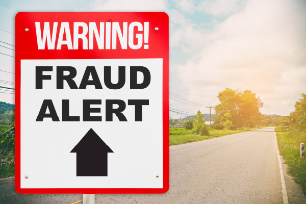 A road sign that says, "Warning! Fraud Alert Ahead," stands in front of a road.