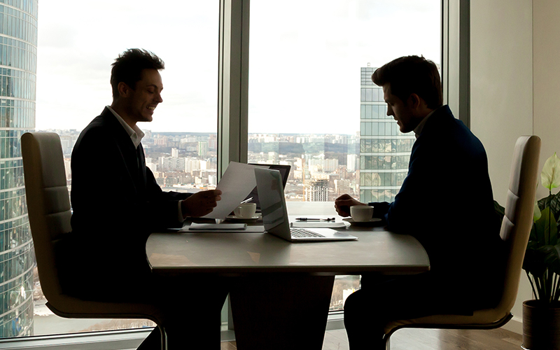 Two silhouetted figures sitting a table in an office, having a discussion.