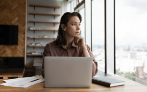 A pensive woman in an office setting at her laptop looking thoughtfully out the window.